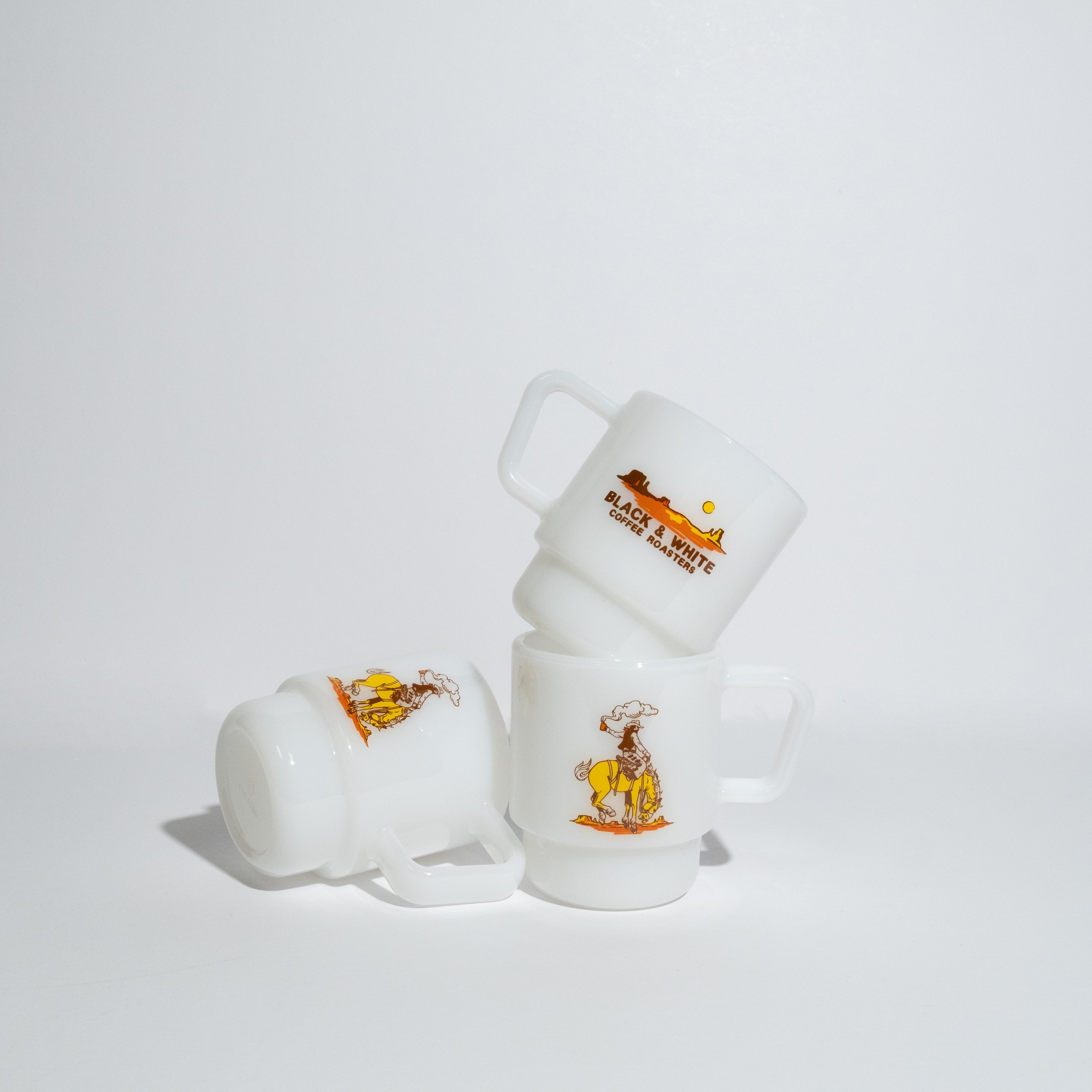 Three small white ceramic mugs, arranged in a stack on a clean white studio background. Each mug features a small printed illustration of a cowboy riding a horse with the text 'Black & White Coffee Roasters' in orange and brown.
