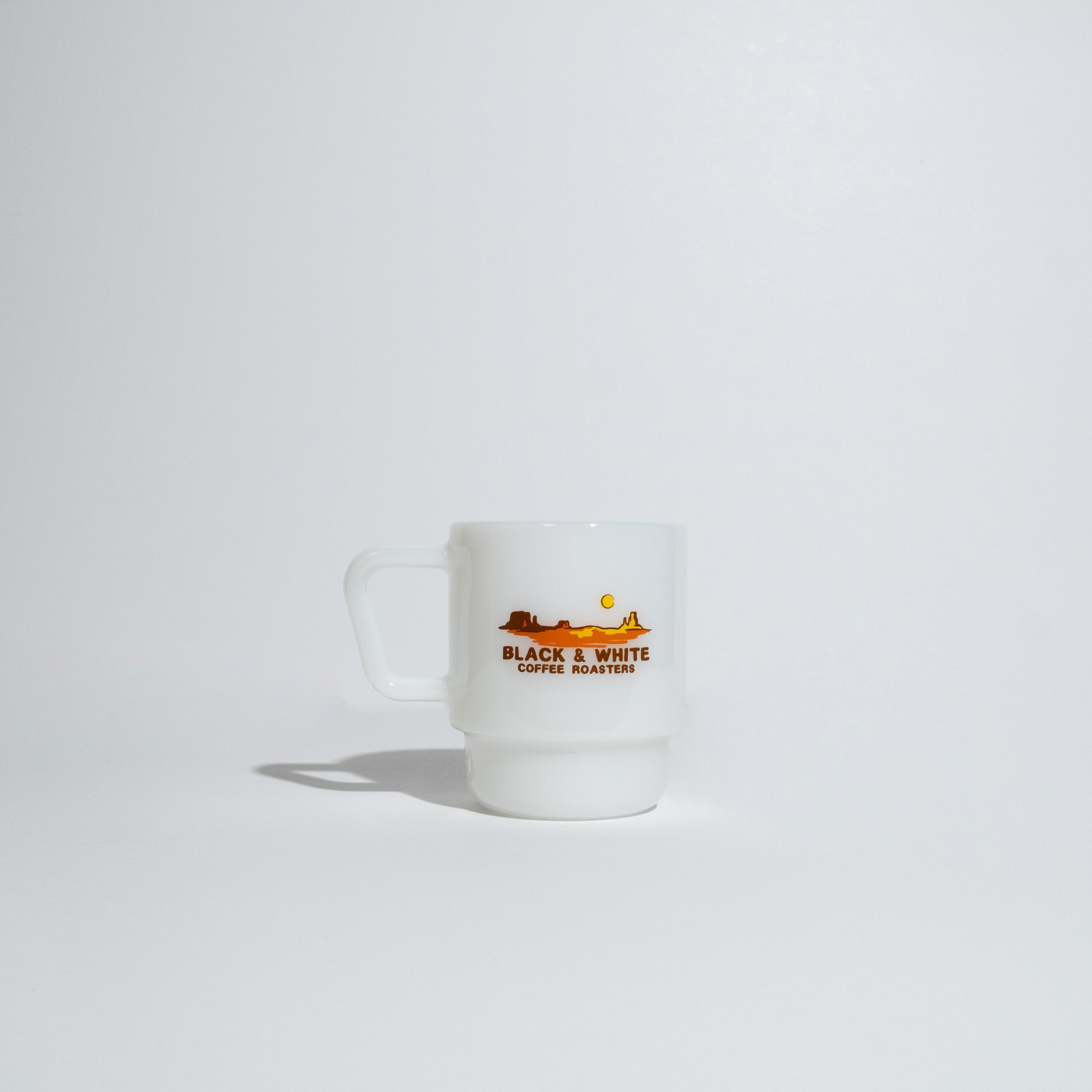 A small white ceramic mug with a handle, featuring a graphic of a sunset over mountains and the text 'BLACK & WHITE COFFEE ROASTERS' on a clean white background.