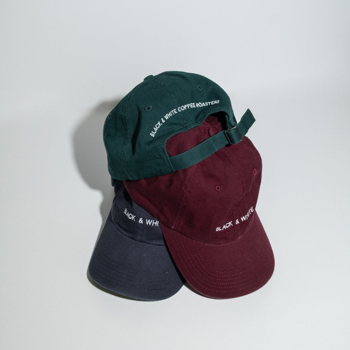 Three baseball caps stacked together on a white surface against a white wall. The top cap is dark green with white embroidered text reading 'BLACK & WHITE COFFEE ROASTERS'; the middle cap is burgundy; the bottom cap is dark navy.