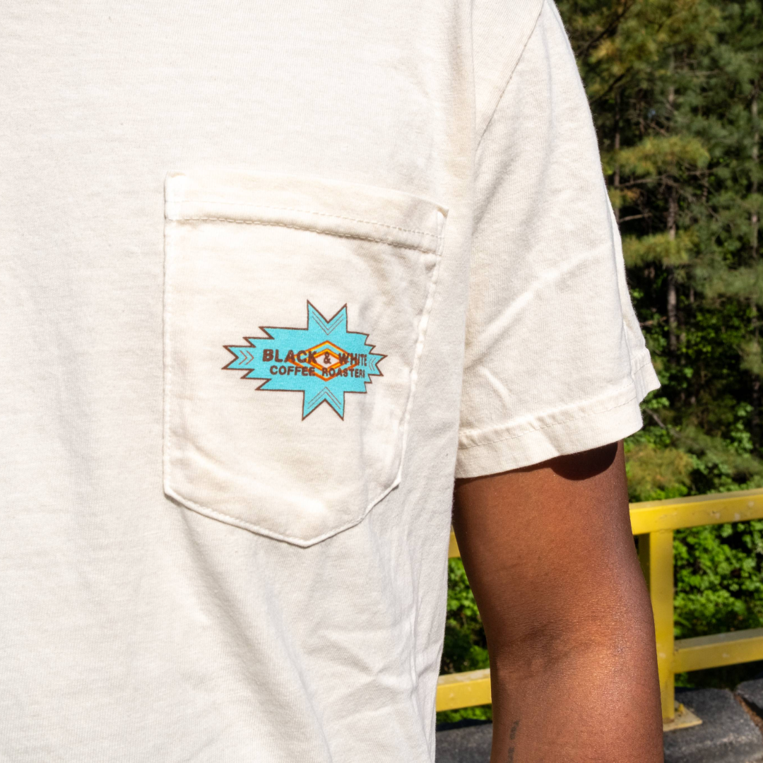 Person wearing Cowboy Pocket Tee with turquoise Black & White Coffee Roasters logo on chest pocket, designed by Tayton Mahone. Outdoors setting visible in background.
