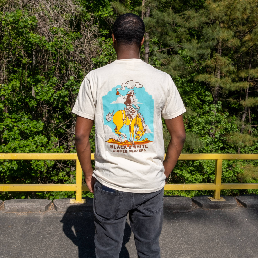 Person wearing Cowboy Pocket Tee, featuring a cowboy graphic with coffee cup, stands near a railing, surrounded by lush greenery. Signature relaxed fit design.