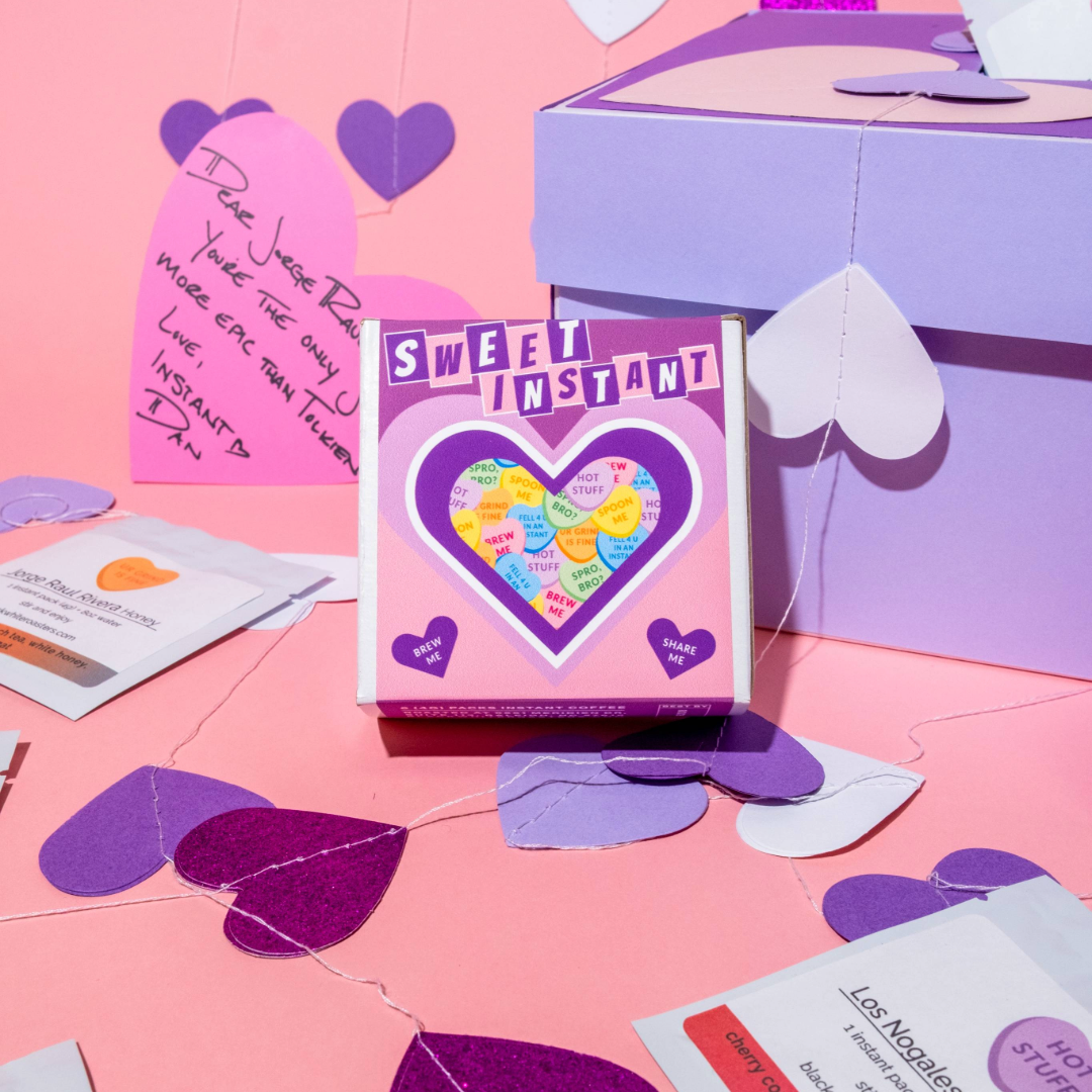 Valentine's Variety Pack - Instant Coffee in a pink box with heart-shaped messages, surrounded by gift boxes, heart garlands, and single-serve coffee packets.