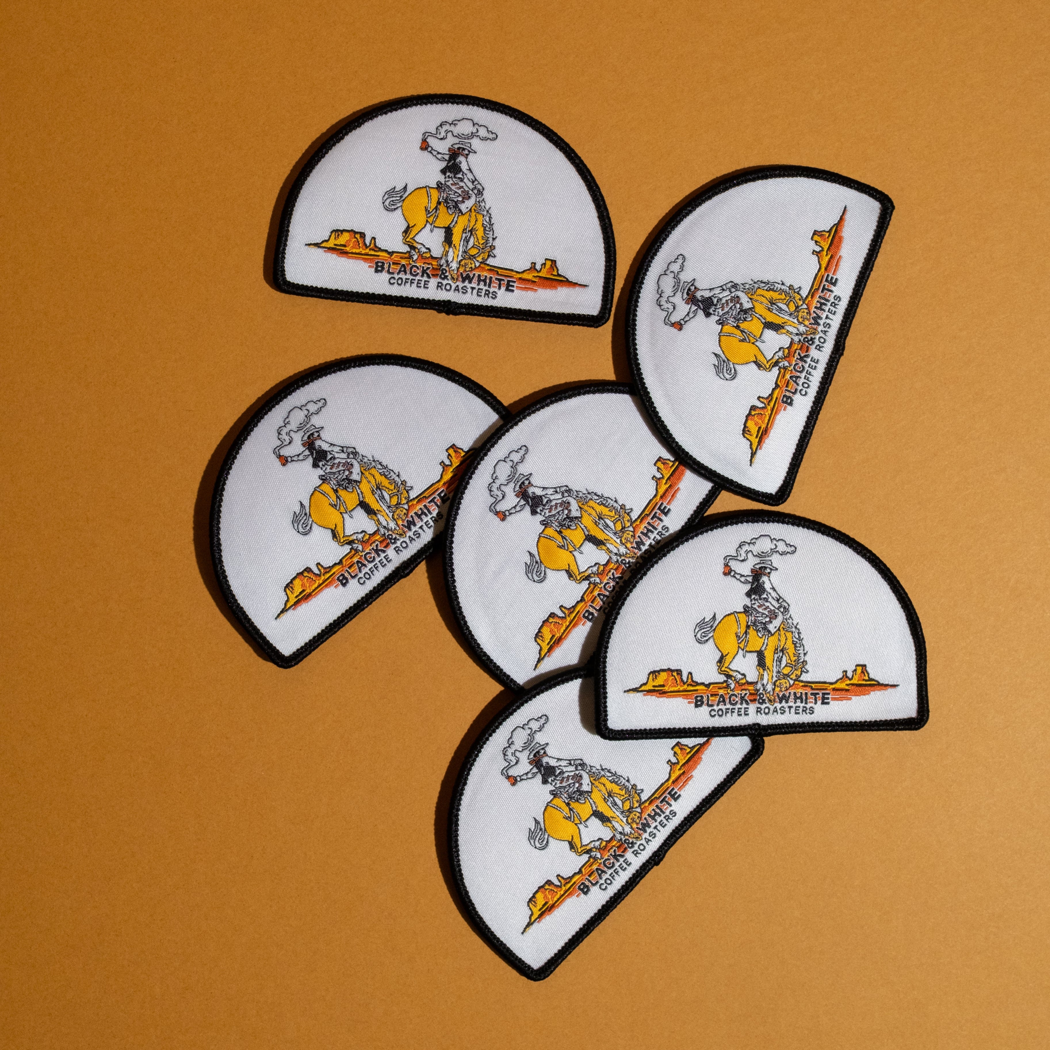 Six white semi-circular patches with black borders, featuring a cartoon cowboy riding a yellow horse and the text 'BLACK & WHITE COFFEE ROASTERS' on an orange background.