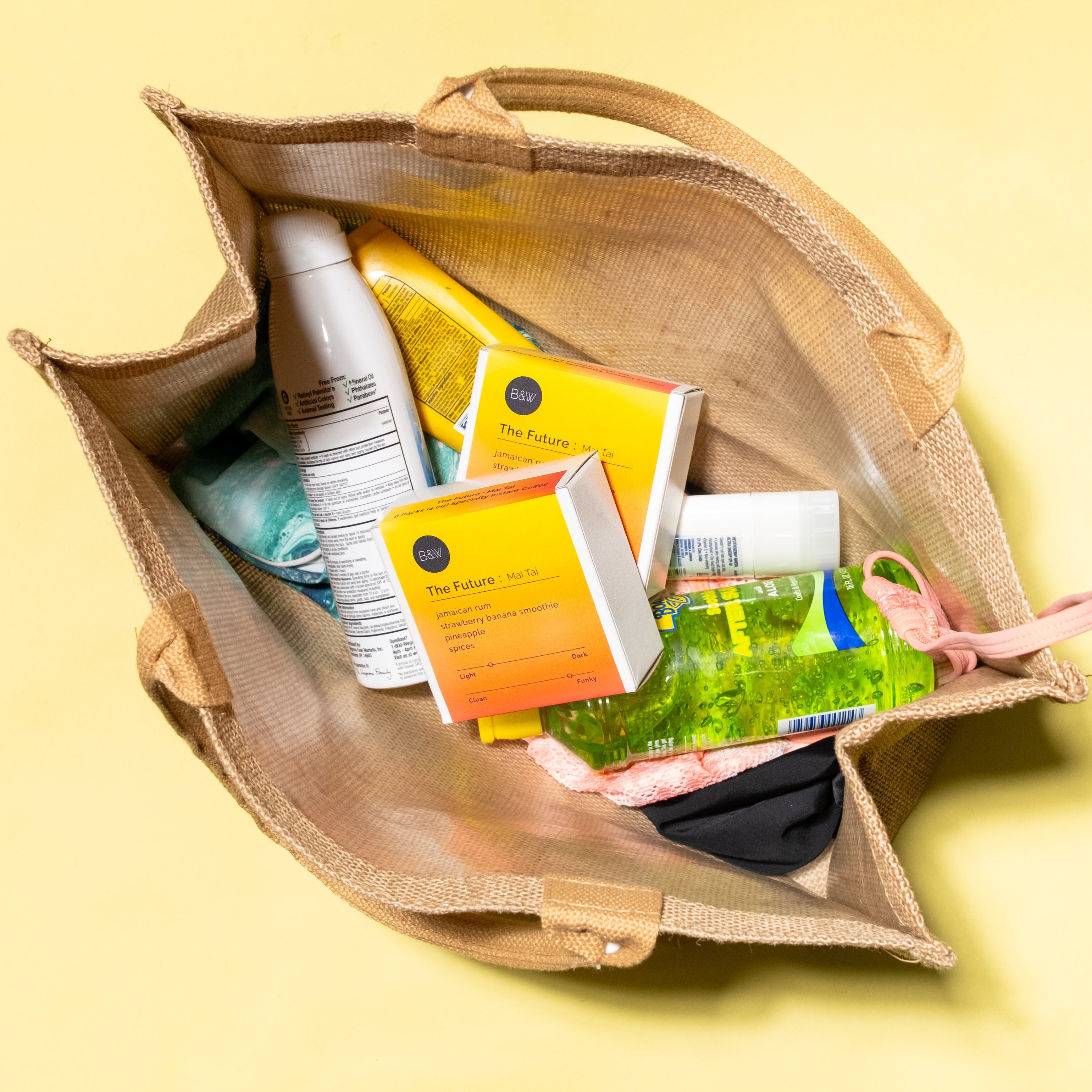 Jute tote with beach essentials, featuring two “The Future: Mai Tai” cocktail-mix boxes, sunscreen, aloe vera gel, green towel, pink goggles, and dark swimwear.