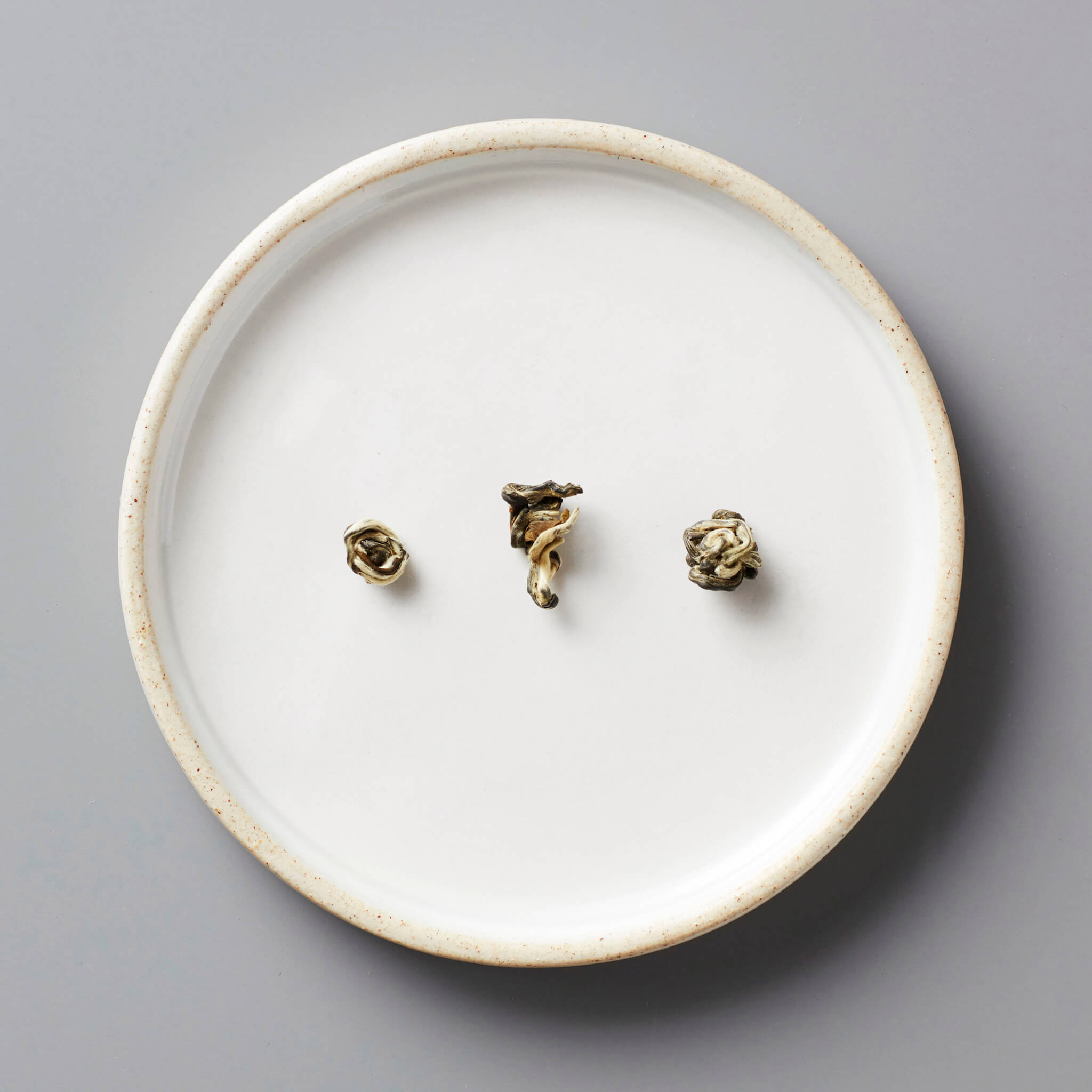Round white ceramic plate with speckled rim holding three tightly rolled tea pearls of Jasmine Bai Hao, a scented green tea from Guangxi, China.